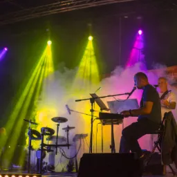 Zespół muzyczny z Bochni w trakcie występu na scenie, oświetlonej kolorowymi reflektorami i wypełnionej dymem. Widoczny klawiszowiec śpiewający do mikrofonu, gitarzysta oraz saksofonista.