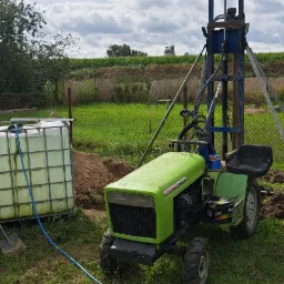 Zielony traktor z wiertnicą na podwórku, obok zbiornik na wodę, w tle pole i pochmurne niebo.