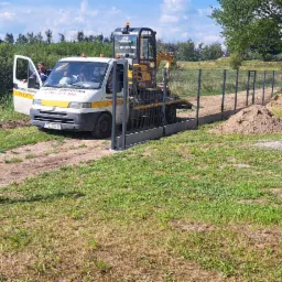 Montaż ogrodzenia panelowego z podmurówką betonową, widoczny samochód pomocy drogowej z mini koparką, świeżo usypane kopce ziemi wzdłuż ogrodzenia.