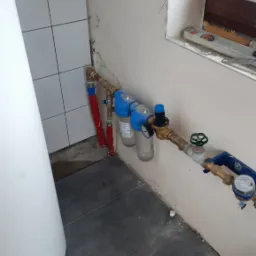 Biały bojler, rury miedziane z filtrami wody i licznikiem, szare płytki podłogowe, ściana z widocznymi śladami prac instalacyjnych.