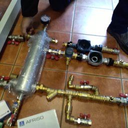 Yurii Hrytsyk - Przygotowanie elementów instalacji: srebrny filtr w folii, mosiężne rury z czerwonymi zaworami, manometr i pudełko AFRISO na brązowych płytkach.