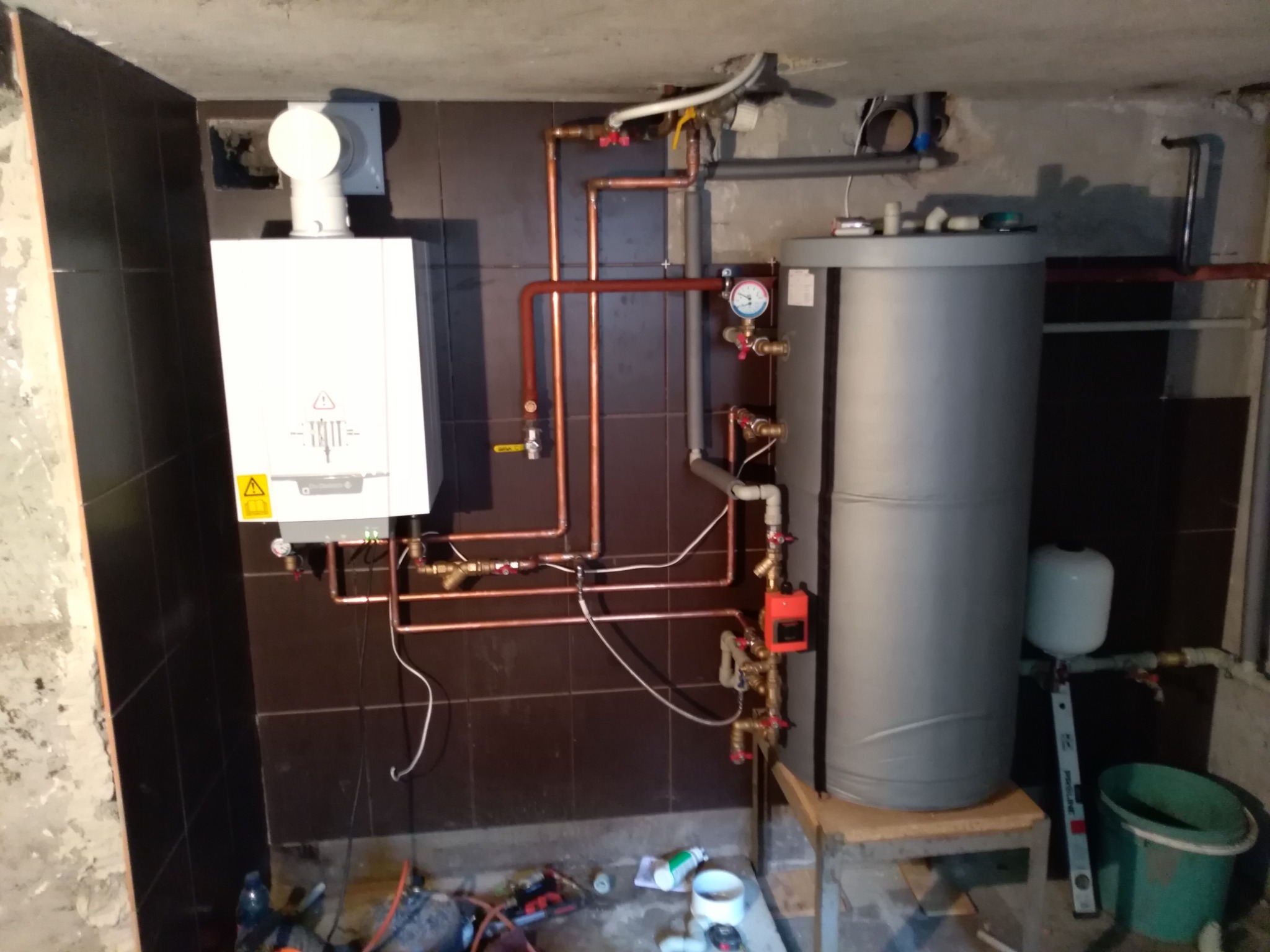 Nowoczesny piec gazowy kondensacyjny z podłączonym zasobnikiem ciepłej wody, instalacja z miedzianych rur, zamontowane na brązowych płytkach, widoczne narzędzia na podłodze w trakcie instalacji.