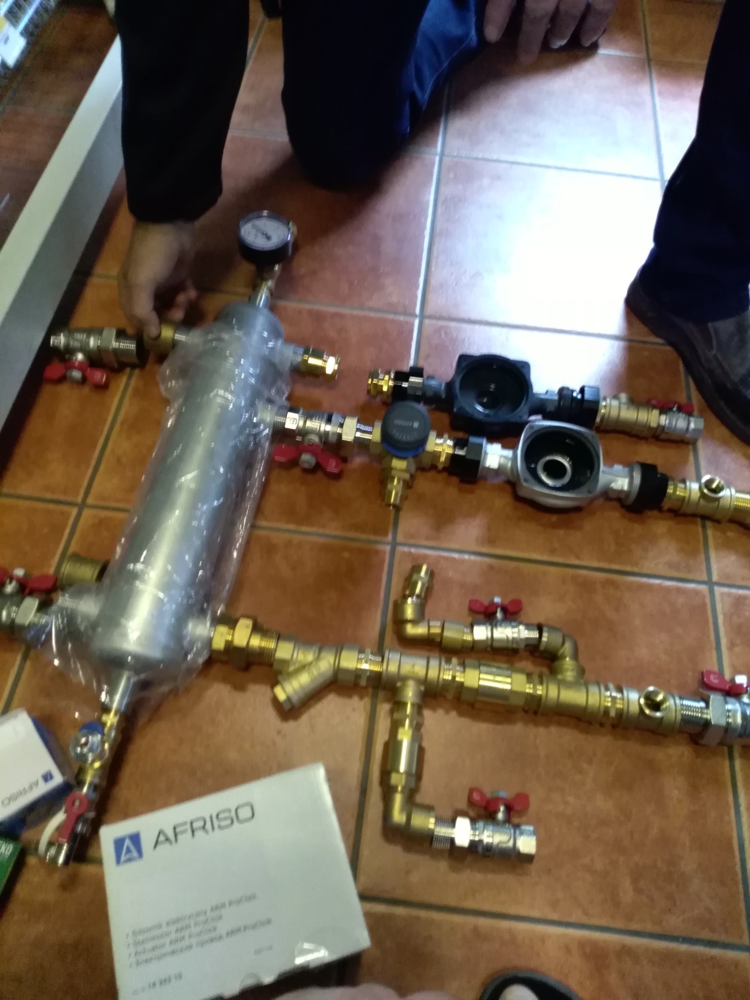 Przygotowanie elementów instalacji: srebrny filtr w folii, mosiężne rury z czerwonymi zaworami, manometr i pudełko AFRISO na brązowych płytkach.
