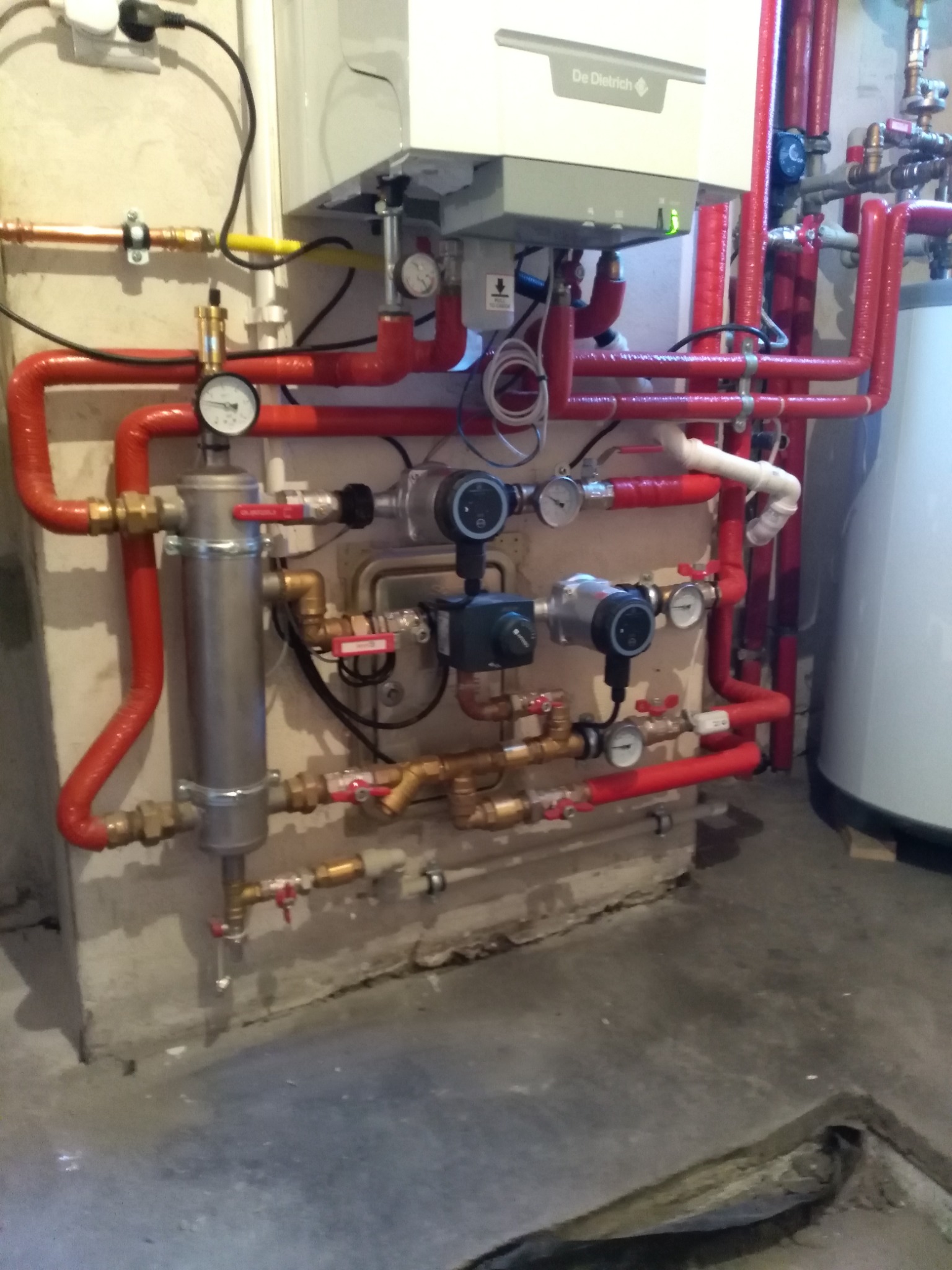 Kotłownia z piecem gazowym De Dietrich, grzejnikiem, pompami obiegowymi, zaworami i rurami w izolacji czerwonej, manometry wskazują ciśnienie w instalacji.