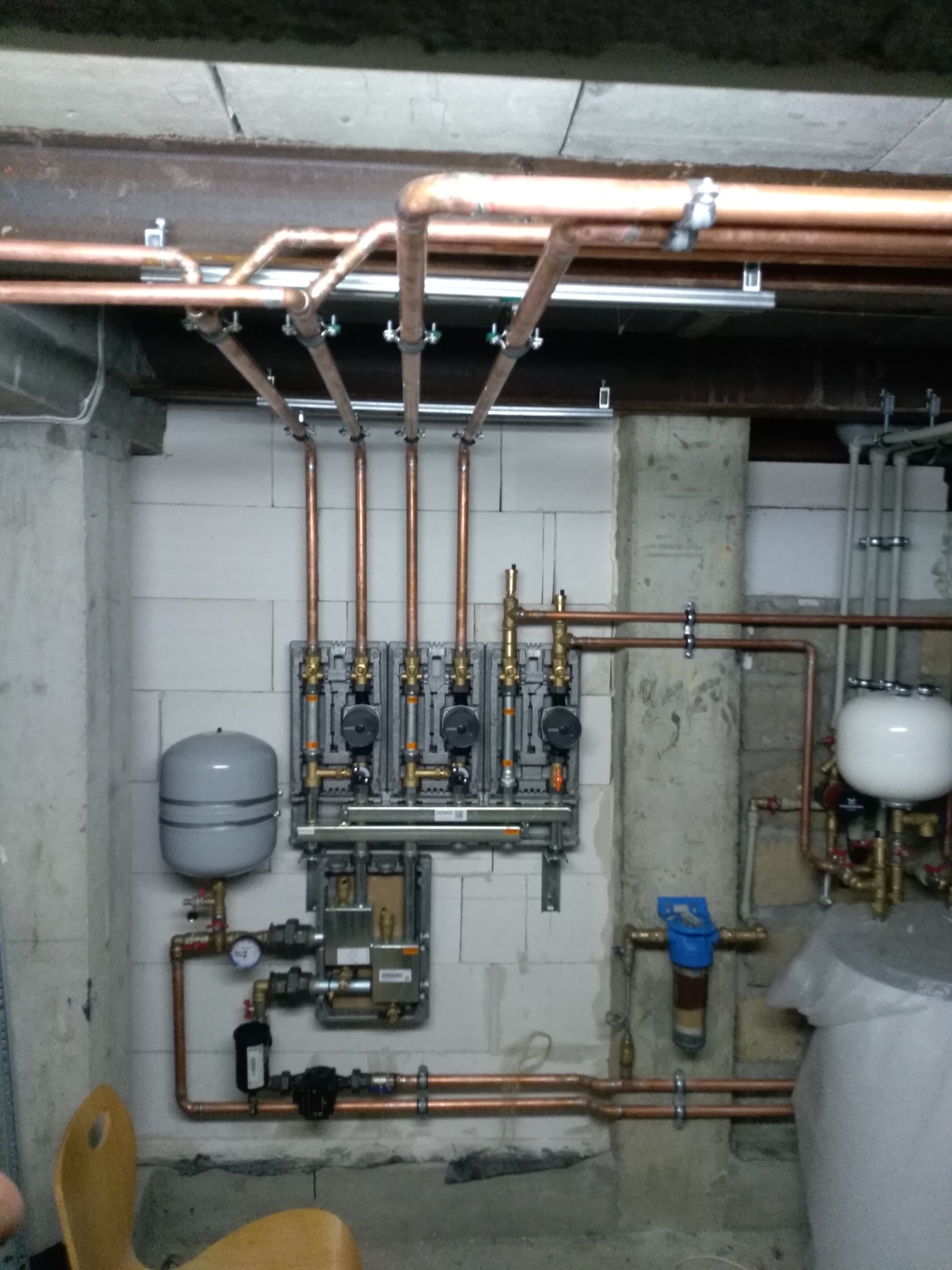 Rozdzielacz ogrzewania podłogowego z widocznymi zaworami, manometrami i rurami miedzianymi, zamontowany na ścianie z betonu komórkowego w surowym wnętrzu.