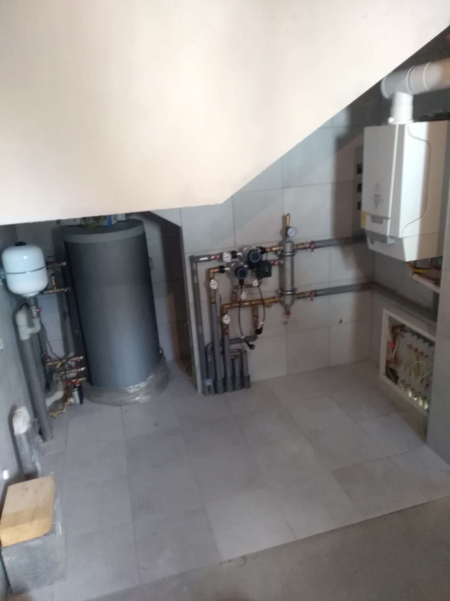 Kotłownia z szarym zbiornikiem akumulacyjnym, białym piecem gazowym kondensacyjnym, rozdzielaczem ogrzewania podłogowego w szafce na ścianie z białych płytek i szarą podłogą z płytek.