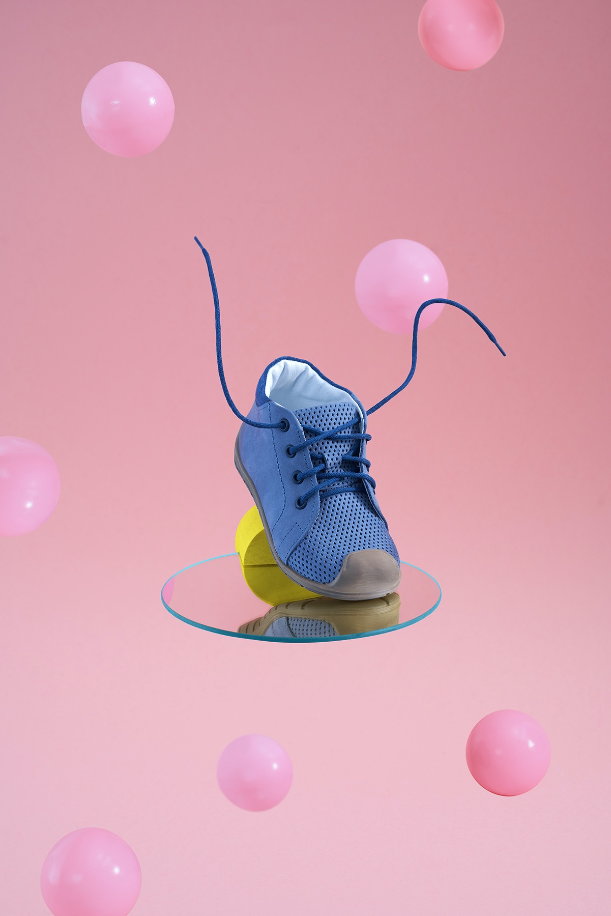 Niebieski, skórzany but dziecięcy z sznurowadłami, umieszczony na okrągłym lustrze i żółtym elemencie, unoszący się na tle różowych kul. Ujęcie z bliska, podkreślające detale wykonania i kolorystykę.