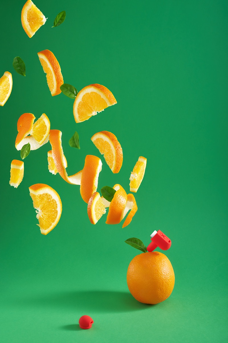 Pomarańcza z czerwonym wyciskaczem, unoszące się plasterki i skórki na zielonym tle, dynamiczna kompozycja.