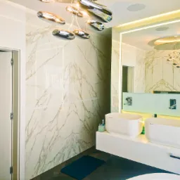 Nowoczesna łazienka z marmurowymi płytkami na ścianach, podwójną umywalką nablatową, podświetlanym lustrem i designerską lampą sufitową w kształcie kropli.