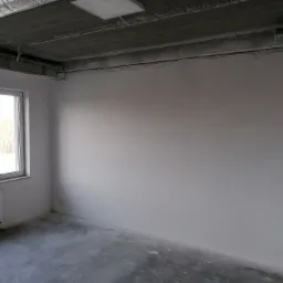 Wnętrze surowego pomieszczenia z widoczną instalacją wentylacyjną pod betonowym stropem, ściany po wstępnym wyrównaniu, przygotowane do dalszych prac wykończeniowych, okno z grzejnikiem po lewej...