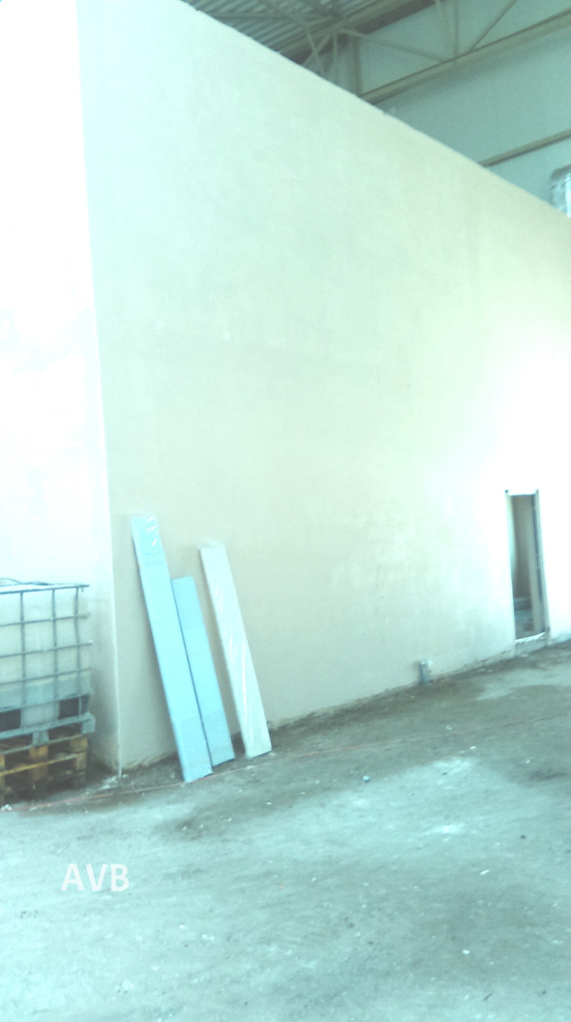 Świeżo otynkowana, jasna ściana w hali produkcyjnej, z widocznymi trzema profilami przy ścianie i paletą z pojemnikiem IBC w rogu, słabe oświetlenie.