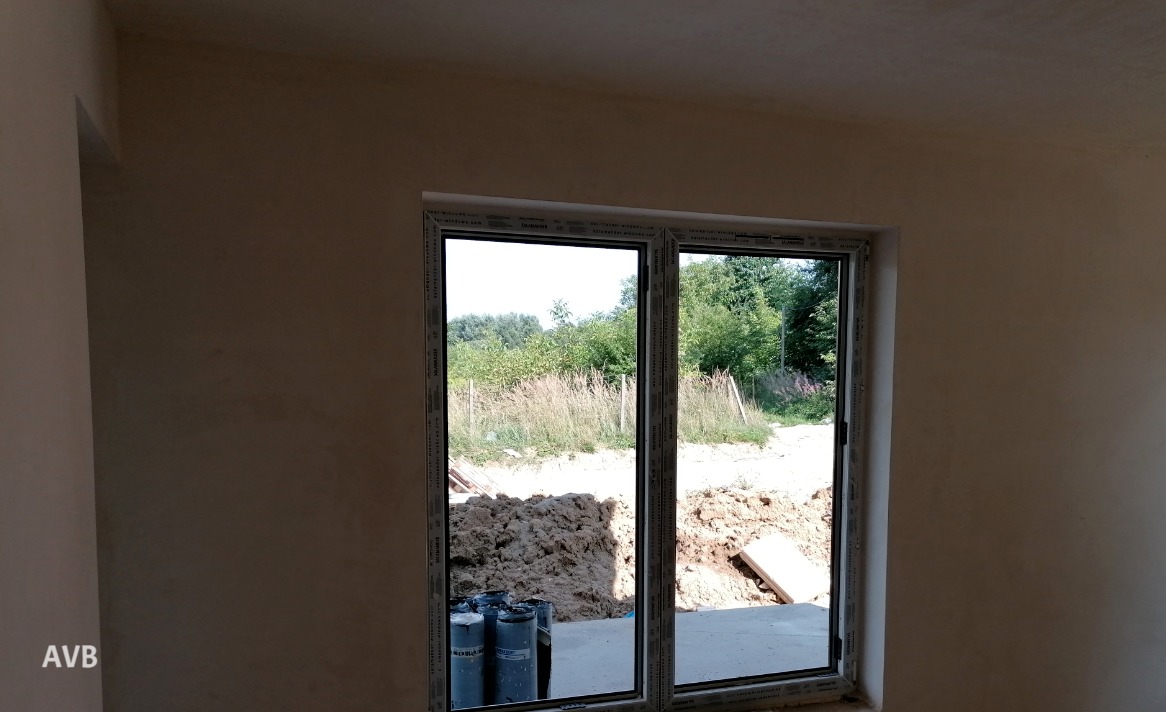 Wnętrze w stanie surowym: ściany po wstępnym przygotowaniu, okno z widokiem na ziemię i zieleń, widoczne butle z gazem.