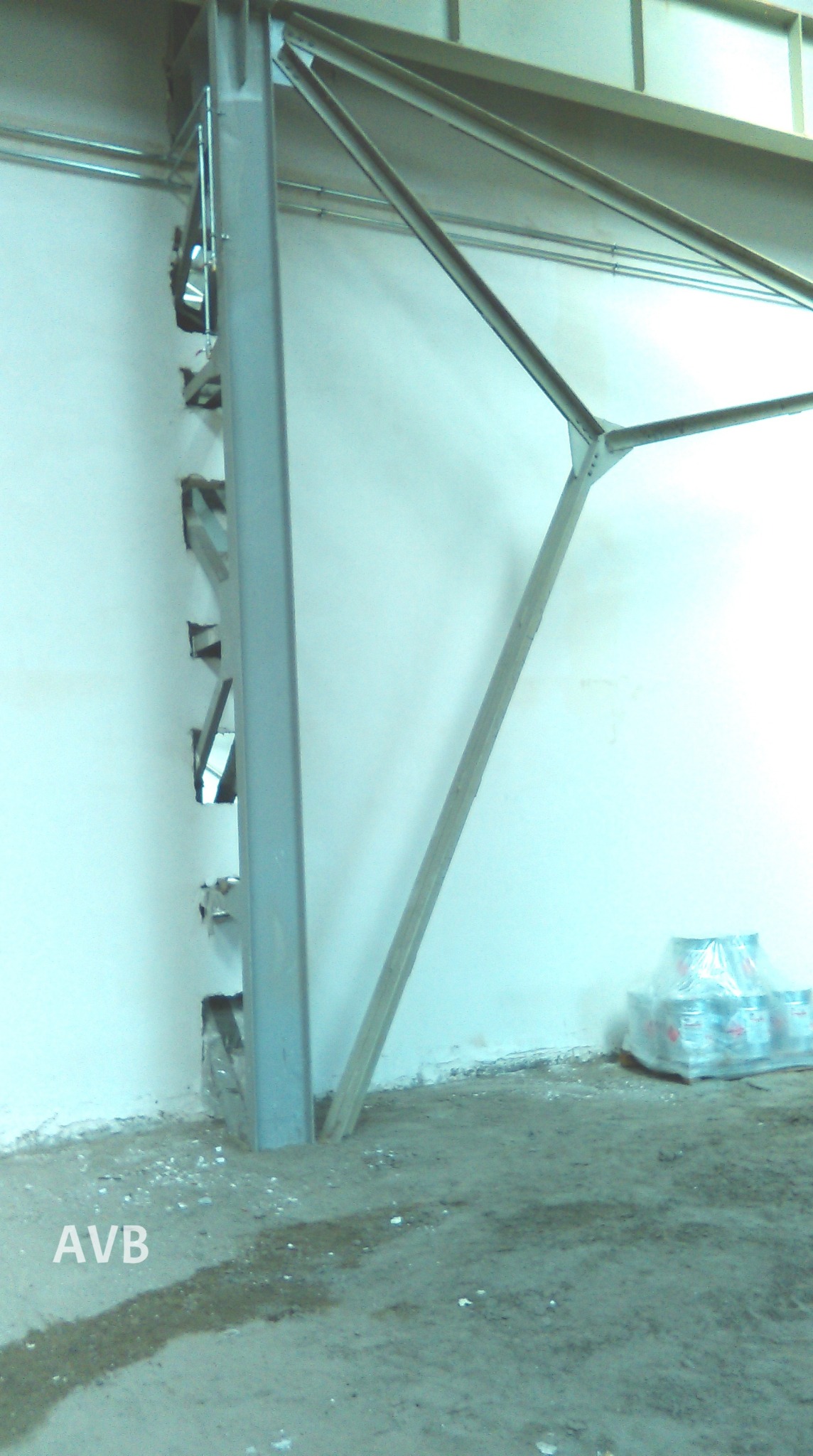 Wnętrze w trakcie prac budowlanych: stalowa konstrukcja wsporcza przy białej ścianie z wyciętymi otworami, zapakowane wiadra z farbą w tle, brudna podłoga.