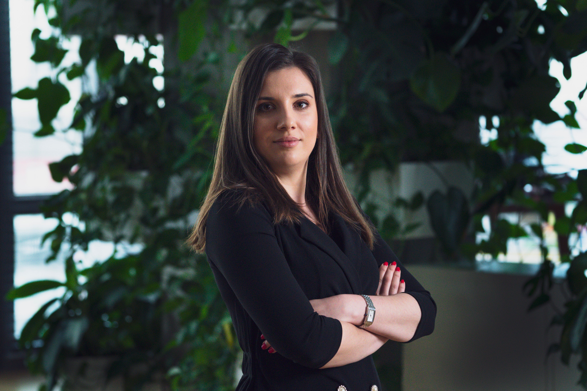 Portret kobiety w czarnej marynarce z założonymi rękami na tle biura z roślinami doniczkowymi.