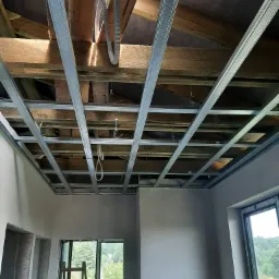 Metalowa konstrukcja podwieszanego sufitu z widocznymi przewodami elektrycznymi, częściowo odsłonięty drewniany strop, okno z widokiem na zieleń, drabina oparta o ścianę w surowym stanie.