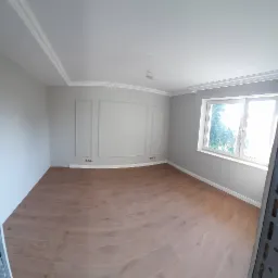 Jasne wnętrze po remoncie: szare ściany z dekoracyjnymi panelami, drewniana podłoga, biały sufit z listwami, duże okno z widokiem na zieleń.