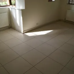 Jasne, kwadratowe płytki podłogowe z ciemnymi fugami w pustym pomieszczeniu z dwoma grzejnikami pod oknami, oświetlone popołudniowym słońcem.