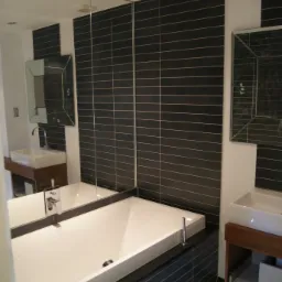 Nowoczesna łazienka z czarnymi płytkami ułożonymi poziomo, białą wanną i umywalkami na drewnianych szafkach, oświetlona punktowymi lampami w suficie i podłodze.