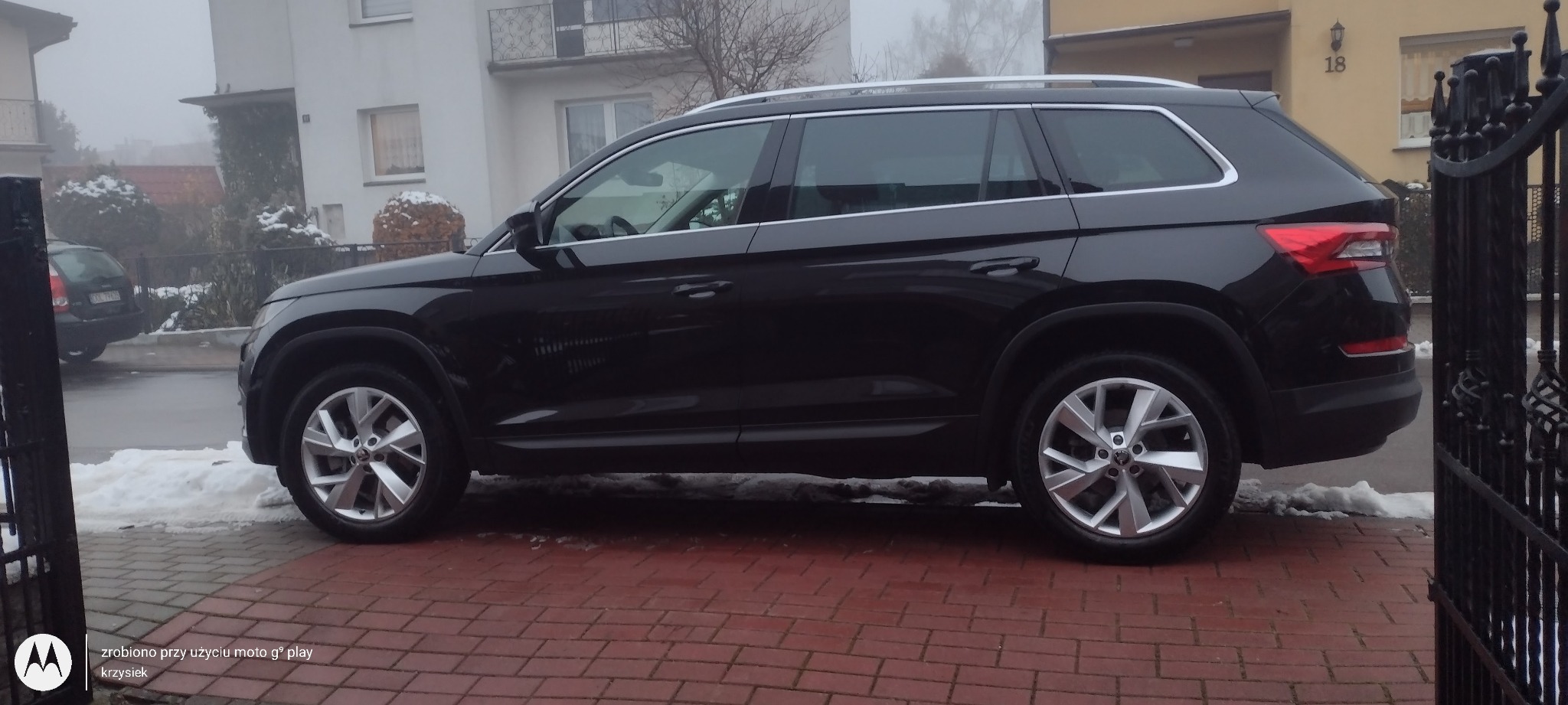 Czarny SUV Skoda Kodiaq zaparkowany na czerwonej kostce brukowej przed domem w pochmurny dzień z resztkami śniegu.