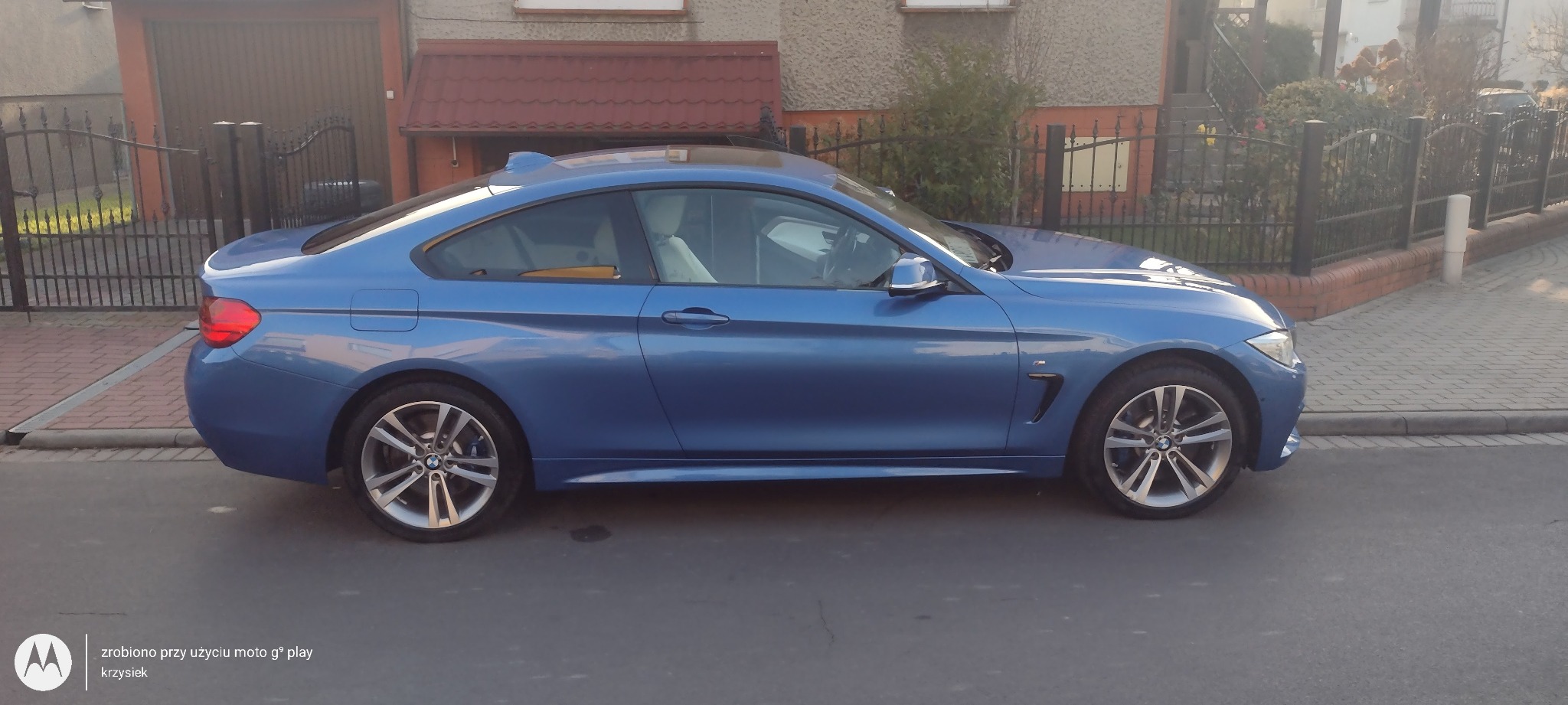 Niebieskie BMW serii 4 Coupe zaparkowane na ulicy przed domem z brązową bramą garażową i żelaznym ogrodzeniem, widok z boku.