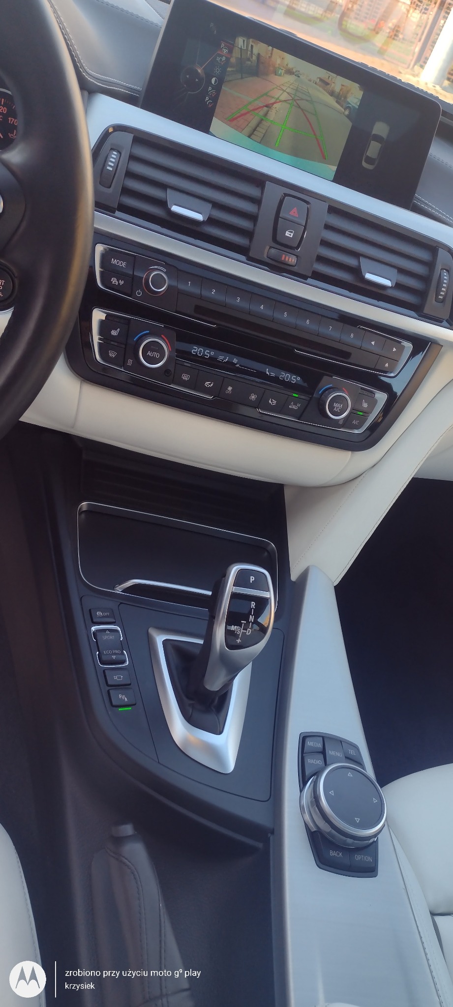 Wnętrze samochodu marki BMW z jasną skórzaną tapicerką, widoczny panel klimatyzacji, automatyczna skrzynia biegów w pozycji parkowania i ekran nawigacji pokazujący obraz z kamery cofania.