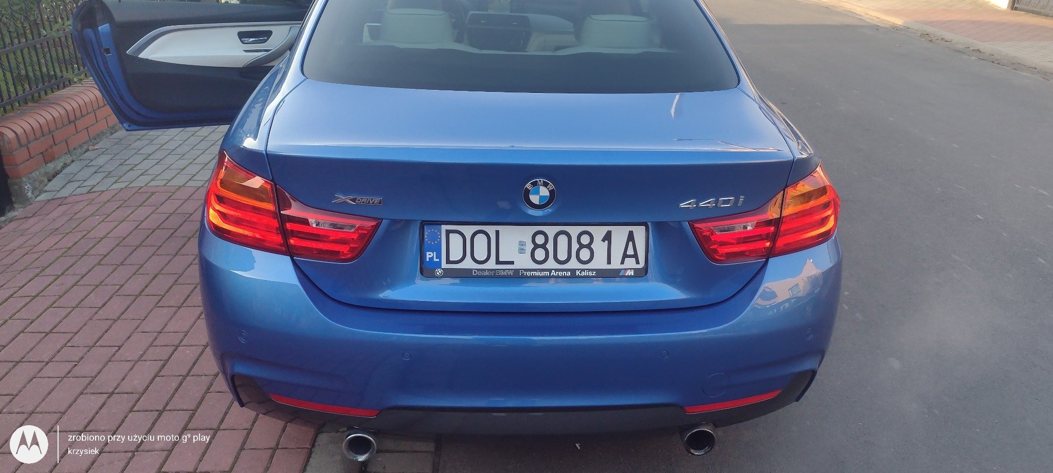 Błękitne BMW serii 4 (440i) z otwartymi drzwiami, widok z tyłu, tablica rejestracyjna DOL 8081A, fragment chodnika z kostki brukowej i asfaltu w tle.