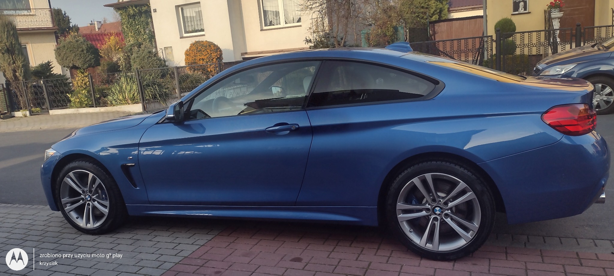 Błękitne BMW serii 4 Coupe zaparkowane na brukowanej ulicy przed domem z zadbanym ogrodem i widocznym innym samochodem w tle.