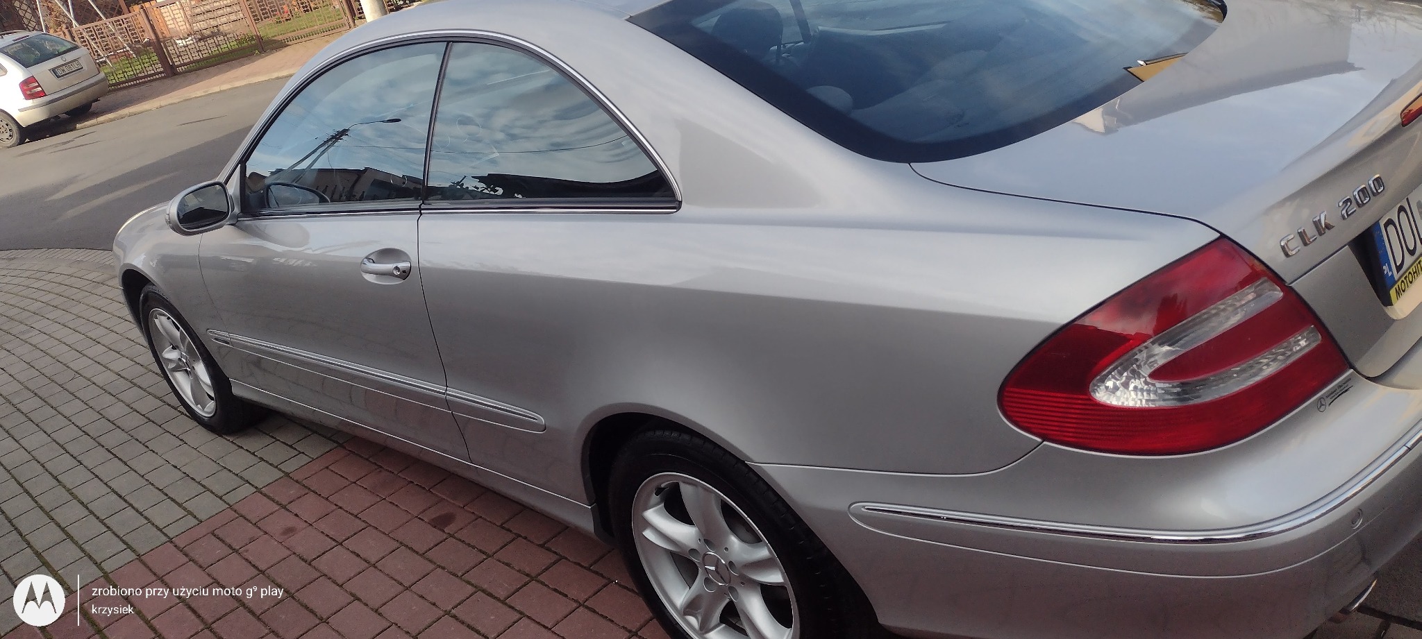 Srebrny Mercedes-Benz CLK 200 na kostce brukowej, widok z boku na tylną część pojazdu, z widocznym odbiciem w szybach