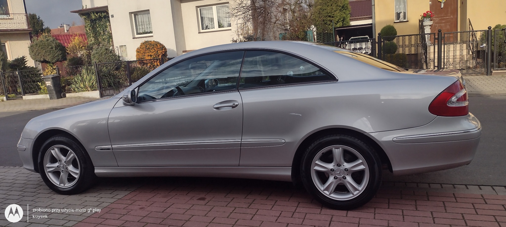 Srebrny Mercedes-Benz CLK zaparkowany na brukowanej ulicy przed domami jednorodzinnymi w słoneczny dzień.