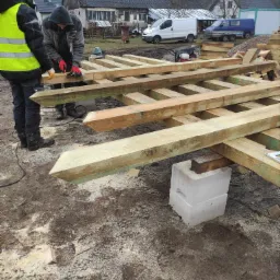 Konstrukcja drewniana z belek na fundamencie z bloczków betonowych, pracownicy w odzieży roboczej przycinają deski, w tle zabudowa wiejska i samochody dostawcze.