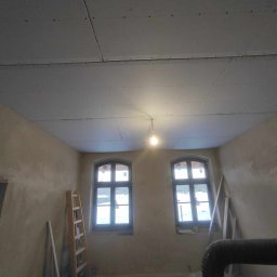 Usługi Ogólnobudowlane Kordek Jan - Wnętrze pokoju w trakcie remontu, sufit pokryty płytami kartonowo-gipsowymi, widoczne dwa okna, drabina oparta o ścianę, ściany w stanie surowym, żarówka zwisająca z sufitu.