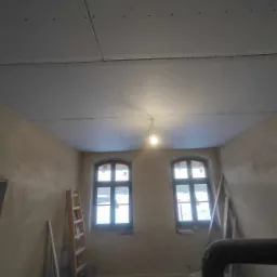 Wnętrze pokoju w trakcie remontu, sufit pokryty płytami kartonowo-gipsowymi, widoczne dwa okna, drabina oparta o ścianę, ściany w stanie surowym, żarówka zwisająca z sufitu.