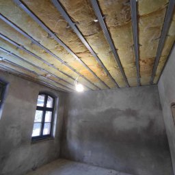 Usługi Ogólnobudowlane Kordek Jan - Wnętrze pokoju w trakcie ocieplania poddasza: widoczne metalowe profile sufitowe z wypełnieniem z wełny mineralnej, surowe szare ściany, okno z czarną ramą i zwisająca żarówka.