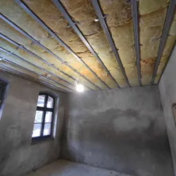 Wnętrze pokoju w trakcie ocieplania poddasza: widoczne metalowe profile sufitowe z wypełnieniem z wełny mineralnej, surowe szare ściany, okno z czarną ramą i zwisająca żarówka.