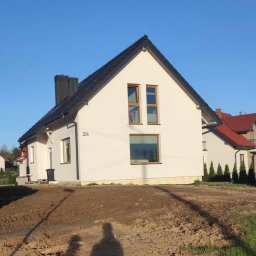 Usługi Ogólnobudowlane Kordek Jan - Nowo wybudowany, otynkowany dom jednorodzinny z ciemną dachówką, z numerem 20 na elewacji, widoczny na tle błękitnego nieba, z niezagospodarowanym terenem przed budynkiem.