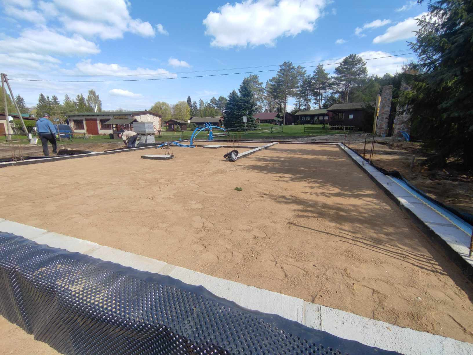 Przygotowany teren pod budowę domu: fundamenty wylewane z betonu, zabezpieczone folią kubełkową, piasek wyrównany, widoczni pracownicy i instalacje.