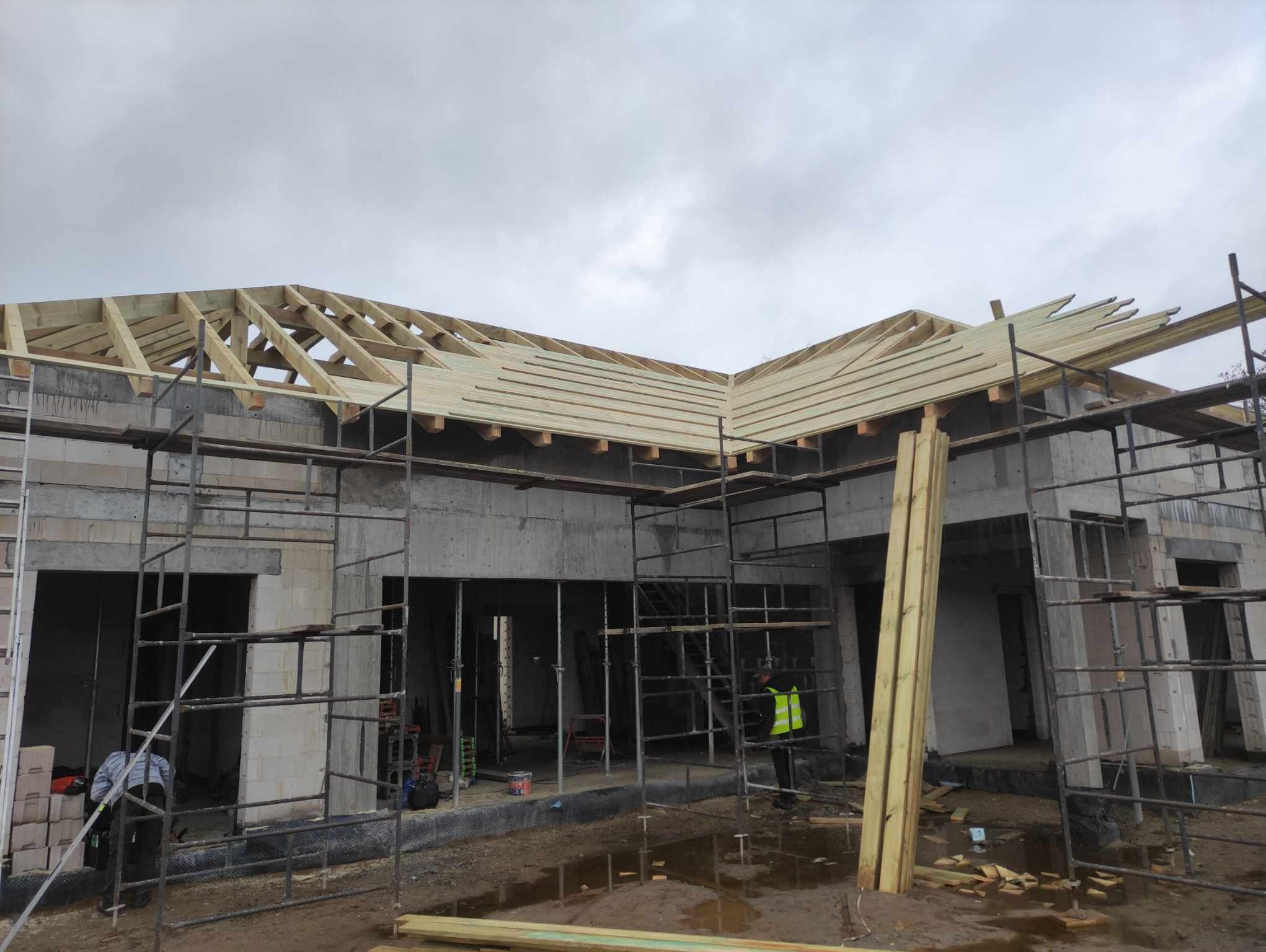 Budowa domu w stanie surowym otwartym: szkielet dachu z drewnianych belek i desek, widoczne rusztowania, pracownik w kamizelce odblaskowej, teren budowy po deszczu.