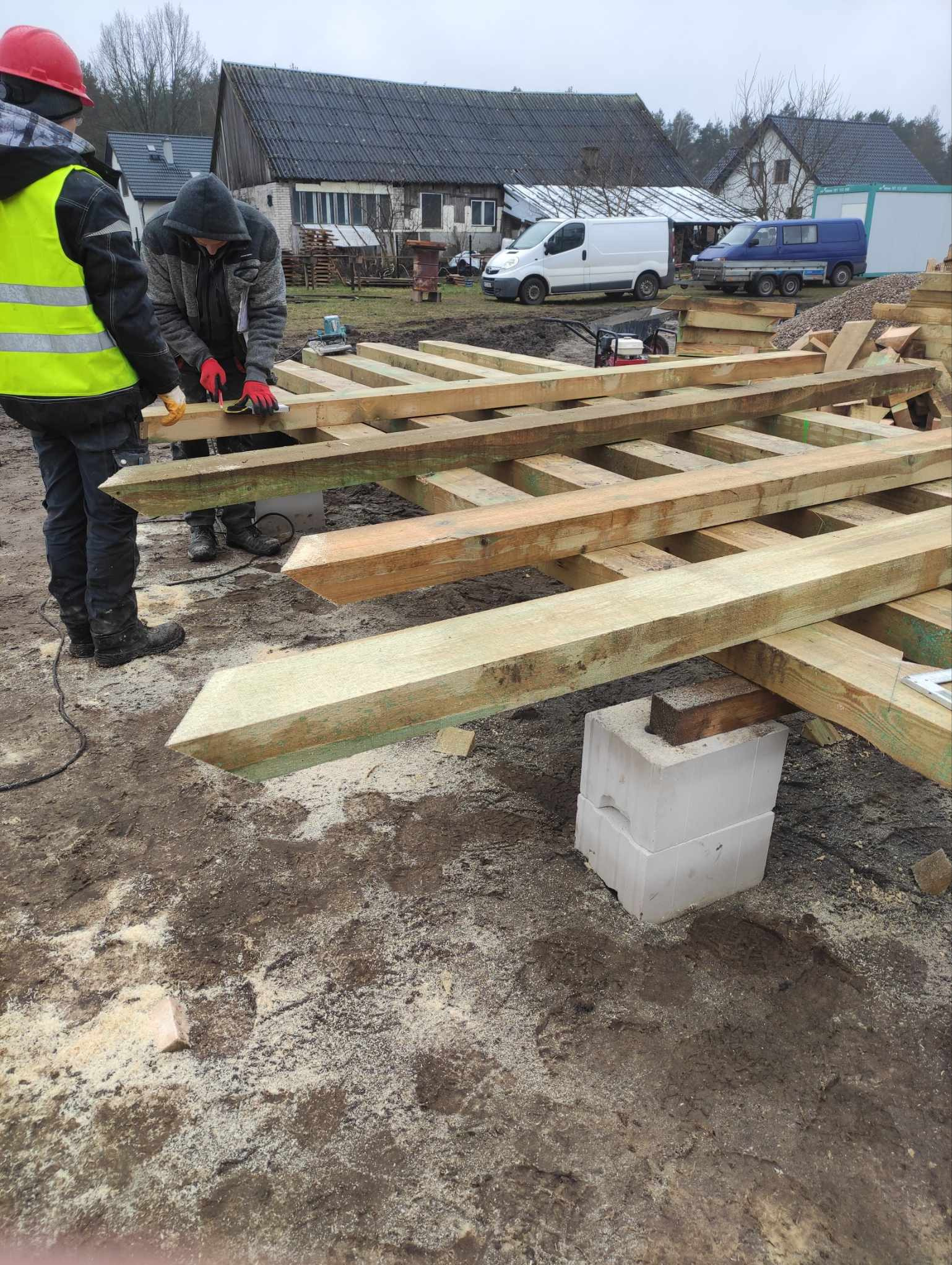Konstrukcja drewniana z belek na fundamencie z bloczków betonowych, pracownicy w odzieży roboczej przycinają deski, w tle zabudowa wiejska i samochody dostawcze.