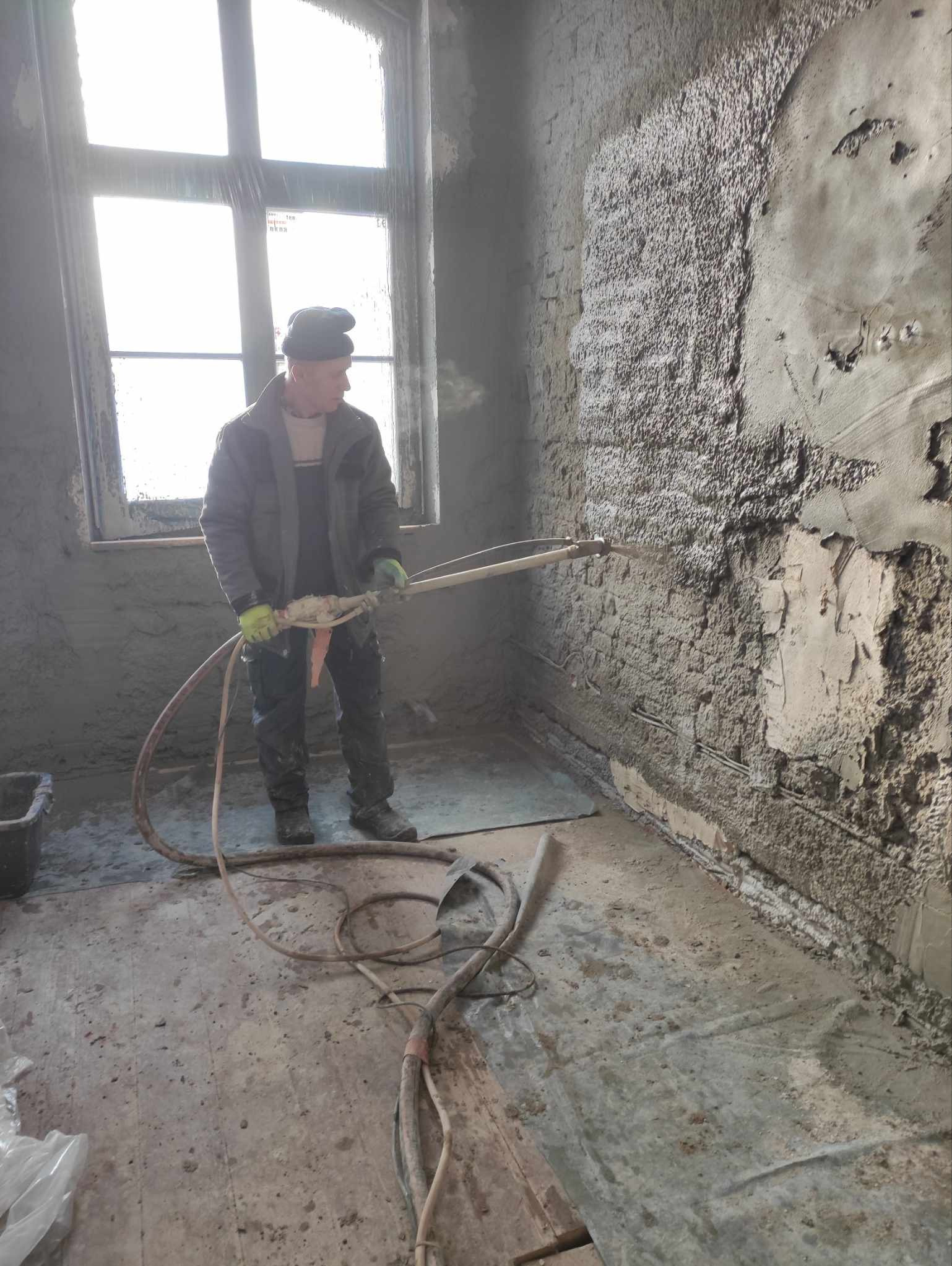 Pracownik w Szczytnie natryskuje tynk na ścianę za pomocą agregatu tynkarskiego w trakcie remontu. Widoczne fragmenty odsłoniętej cegły i instalacji elektrycznej.