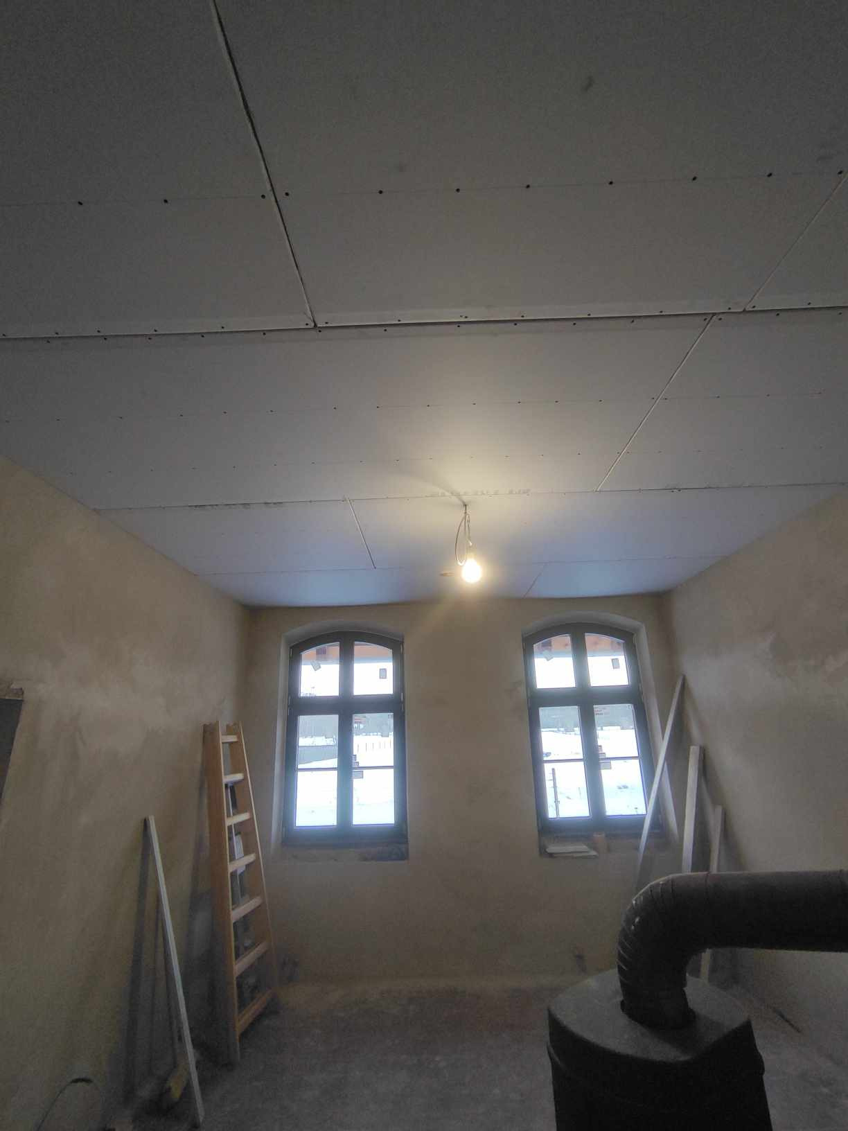 Wnętrze pokoju w trakcie remontu, sufit pokryty płytami kartonowo-gipsowymi, widoczne dwa okna, drabina oparta o ścianę, ściany w stanie surowym, żarówka zwisająca z sufitu.