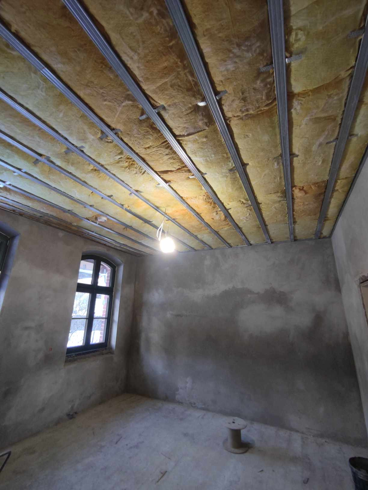 Wnętrze pokoju w trakcie ocieplania poddasza: widoczne metalowe profile sufitowe z wypełnieniem z wełny mineralnej, surowe szare ściany, okno z czarną ramą i zwisająca żarówka.