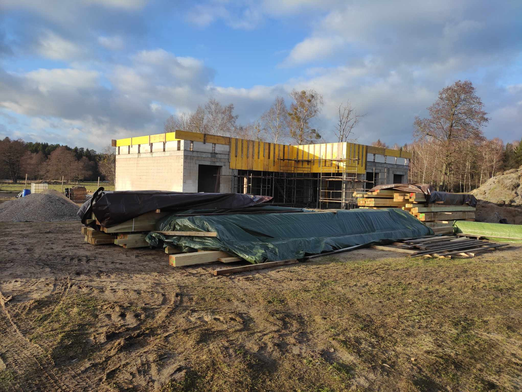 Budowa domu murowanego w trakcie realizacji: widoczne ściany z bloczków, żółte szalunki na stropie, rusztowania, składowane drewno budowlane i materiały przykryte folią, ziemisty teren budowy...