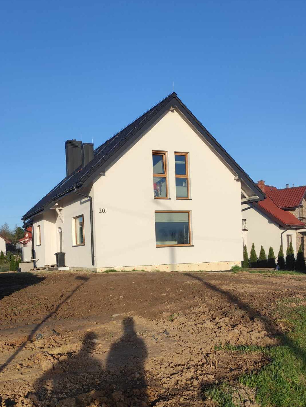 Nowo wybudowany, otynkowany dom jednorodzinny z ciemną dachówką, z numerem 20 na elewacji, widoczny na tle błękitnego nieba, z niezagospodarowanym terenem przed budynkiem.
