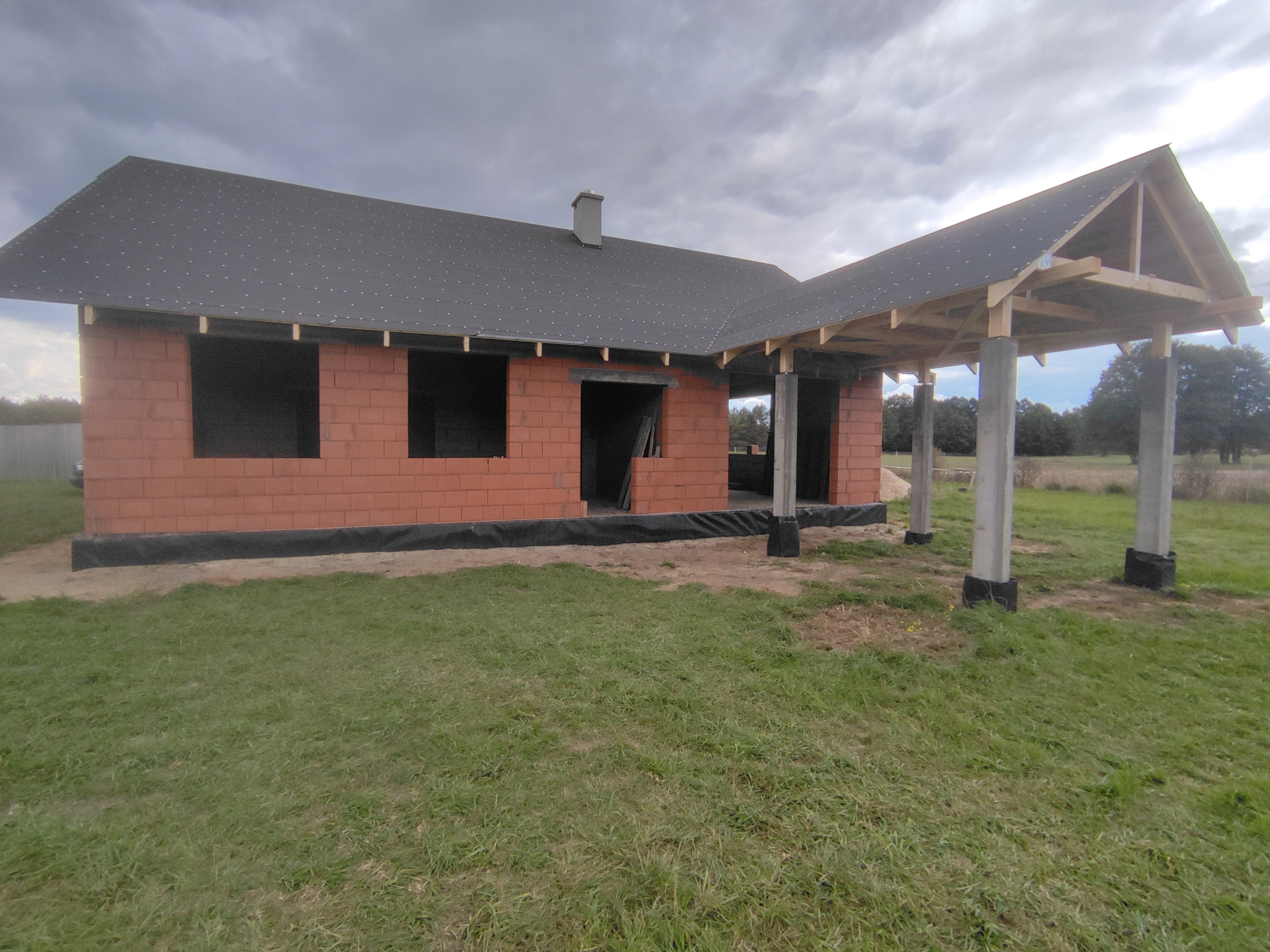 Surowy stan domu jednorodzinnego z czerwonej cegły z dachem pokrytym papą, widoczne betonowe słupy podtrzymujące drewnianą konstrukcję zadaszenia, w tle zielona trawa i pochmurne niebo.