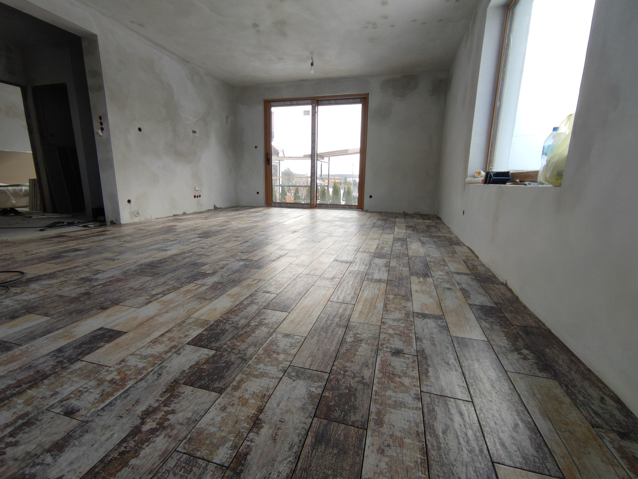 Surowe wnętrze z drewnopodobną podłogą, ściany w stanie deweloperskim, duże przesuwne okno balkonowe z widokiem na zewnątrz, na parapecie butelka z płynem i wałek malarski.