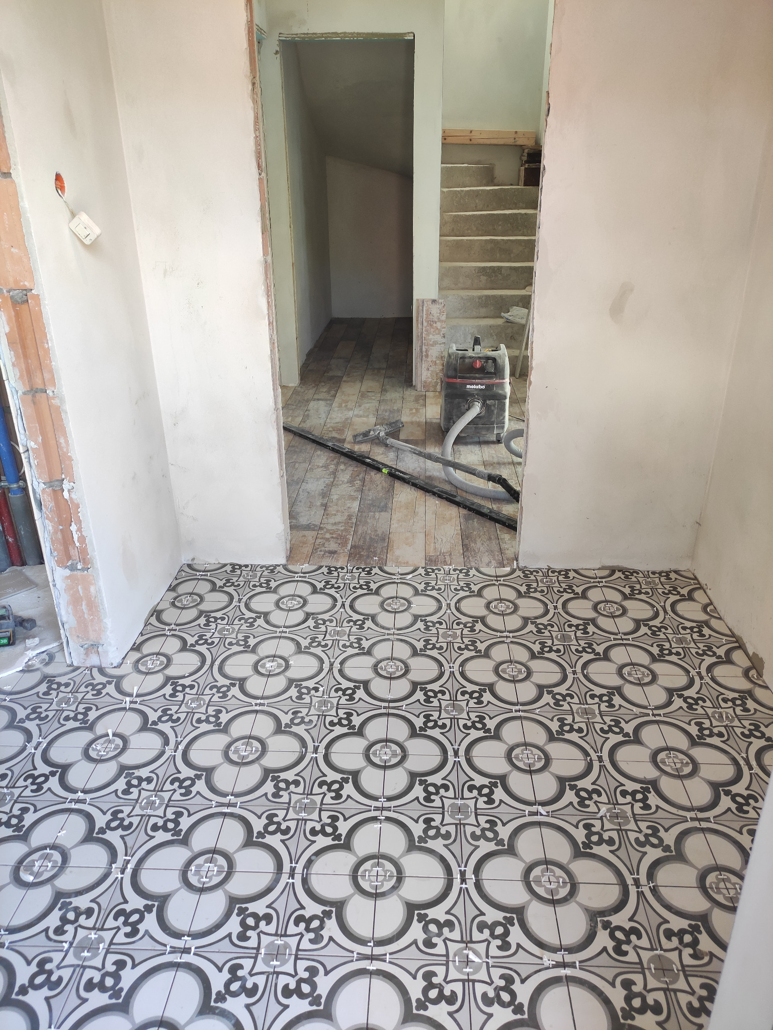 Wnętrze w trakcie remontu: świeżo położona podłoga z płytek w kwiatowy wzór, widoczny odkurzacz przemysłowy Metabo, surowe ściany w trakcie wykańczania i schody w tle.