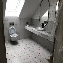 Nowoczesna łazienka na poddaszu z płytkami lastryko na podłodze i blacie, białe pionowe płytki na ścianach, owalne lustra w czarnych ramach i toaleta pod oknem dachowym.