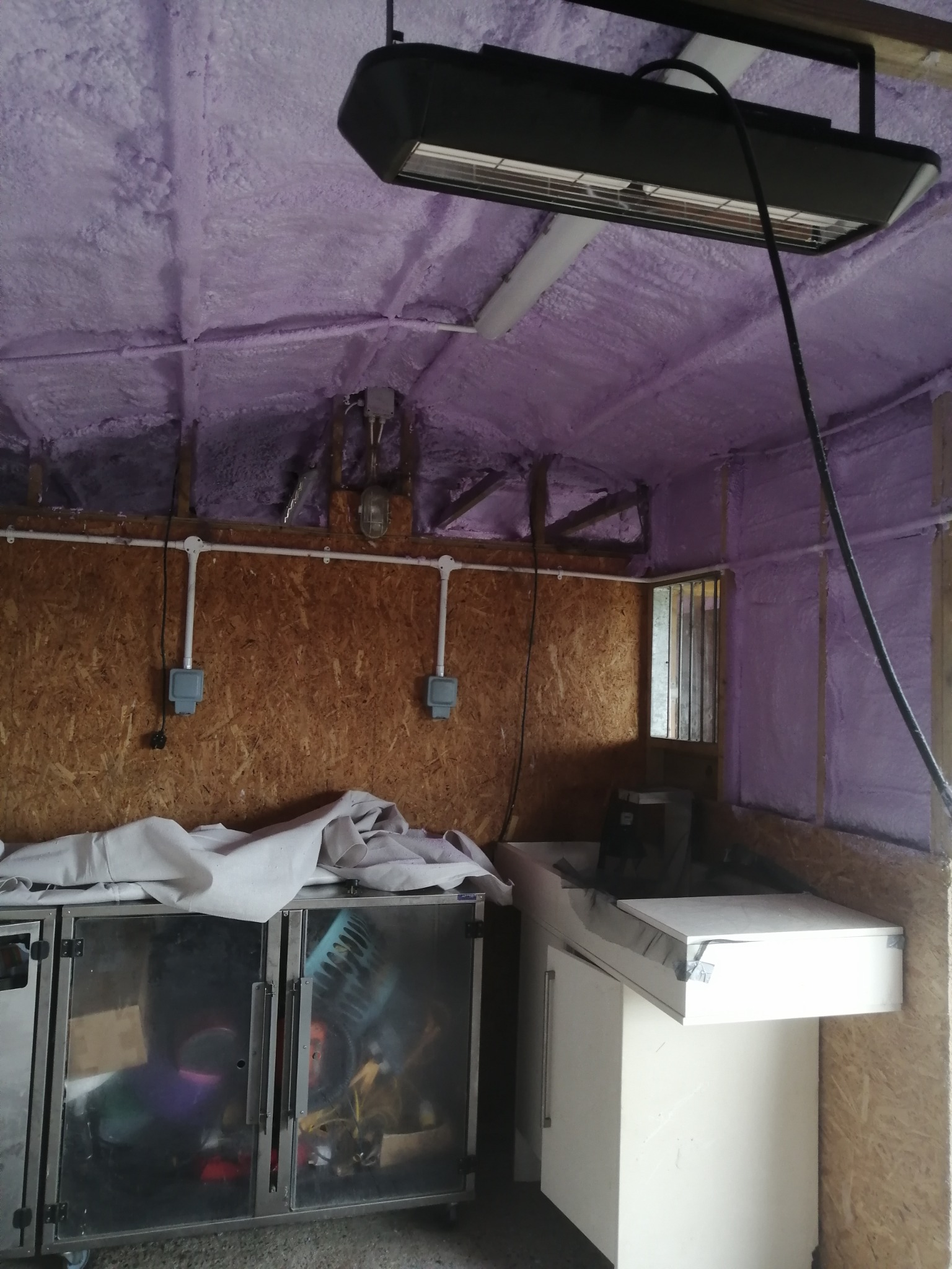Poddasze garażu po natrysku pianki poliuretanowej w kolorze fioletowym, widoczne instalacje elektryczne, lampa grzewcza pod sufitem oraz meble warsztatowe w dolnej części kadru.