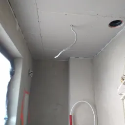 Montaż sufitu podwieszanego z płyt gipsowo-kartonowych w łazience, widoczne przewody elektryczne i rury instalacji wodnej, ściany przygotowane do dalszych prac wykończeniowych.