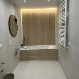 Nowoczesna łazienka w trakcie wykańczania: beżowe płytki, wanna obudowana panelami, okrągłe lustro, grzejnik drabinkowy, widoczne niedokończone przyłącza i wiadro z zaprawą.